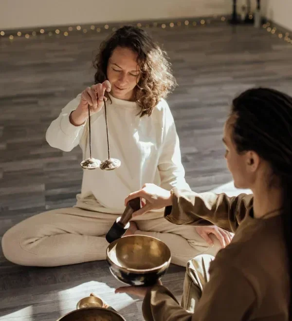 This images shows two girls are sitting on the brown coloured floor, having some cooking utensils in their hands, one girl is engaged in serving food and another is observing the another tool , like try to find how it works, a morning light comes from background.