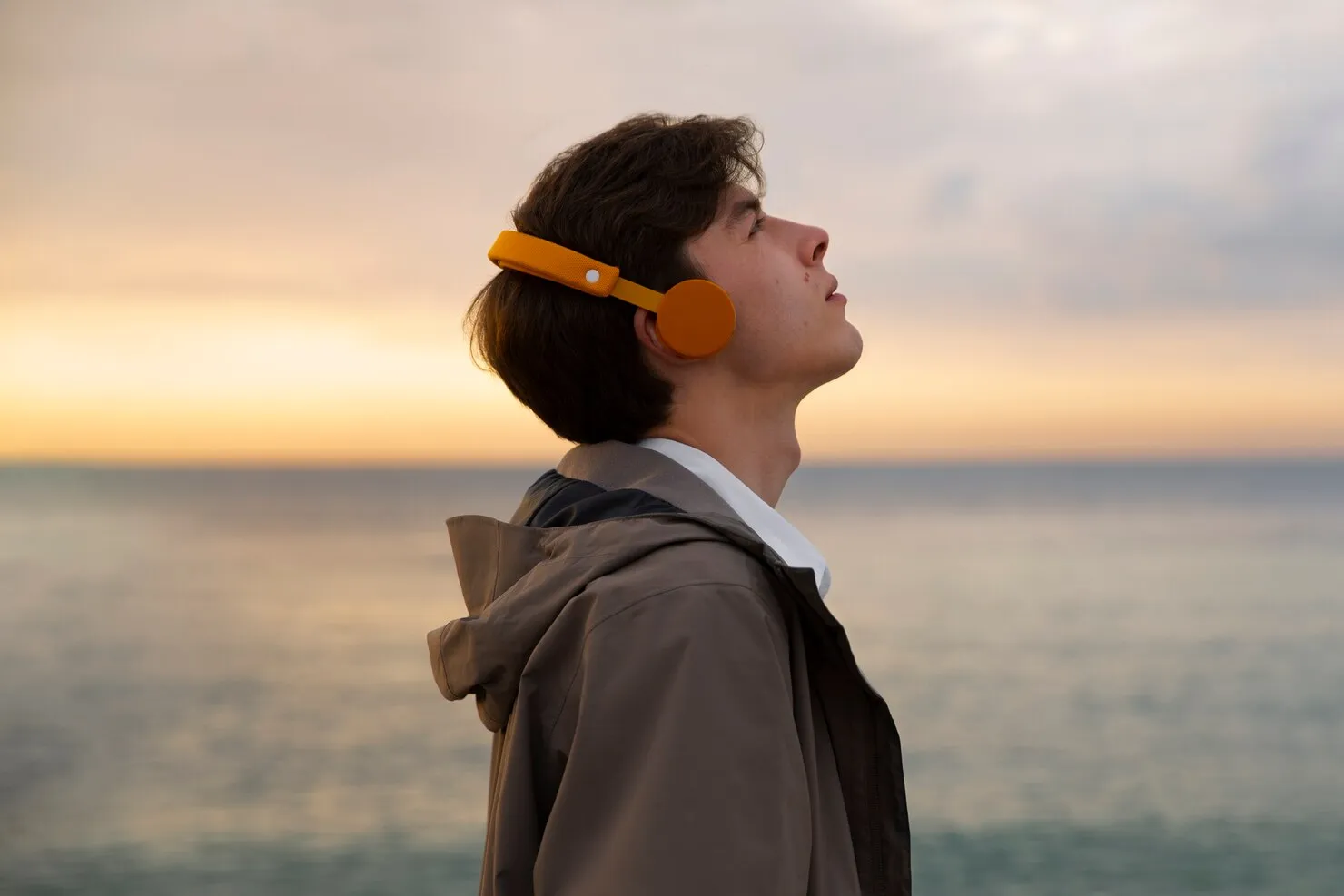 A boy with headphones, looked too much confused view towards the sky , a pretty weather and ocean is present behind him.