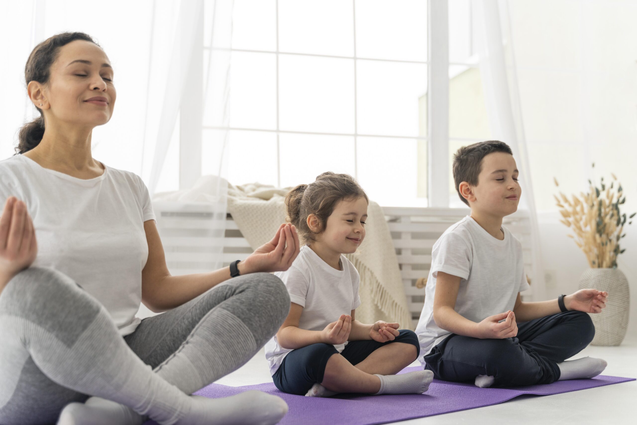 Scene appears a femail with two kids doing meditation with smily face sitted on the blue coloured mat. Having a window , white coloured curtains and plant pots are present as the background.