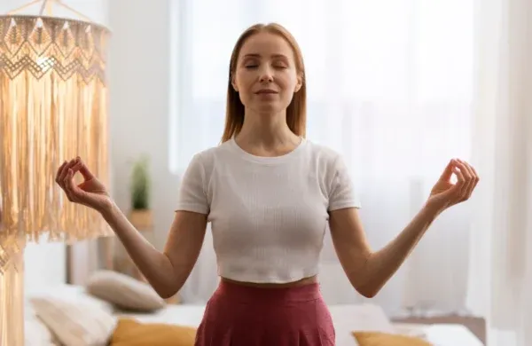 A depiction of upper half of foreign lady doing Nostril breathing in a room , having some white curtains that cover the window,sofa and pillows as the background portion.