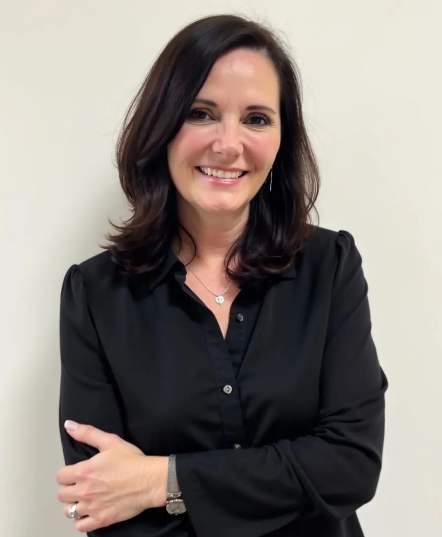 Wendy Byrd is standing with a smile, wearing a black shirt and folding her hands, set against a cream-patterned background.