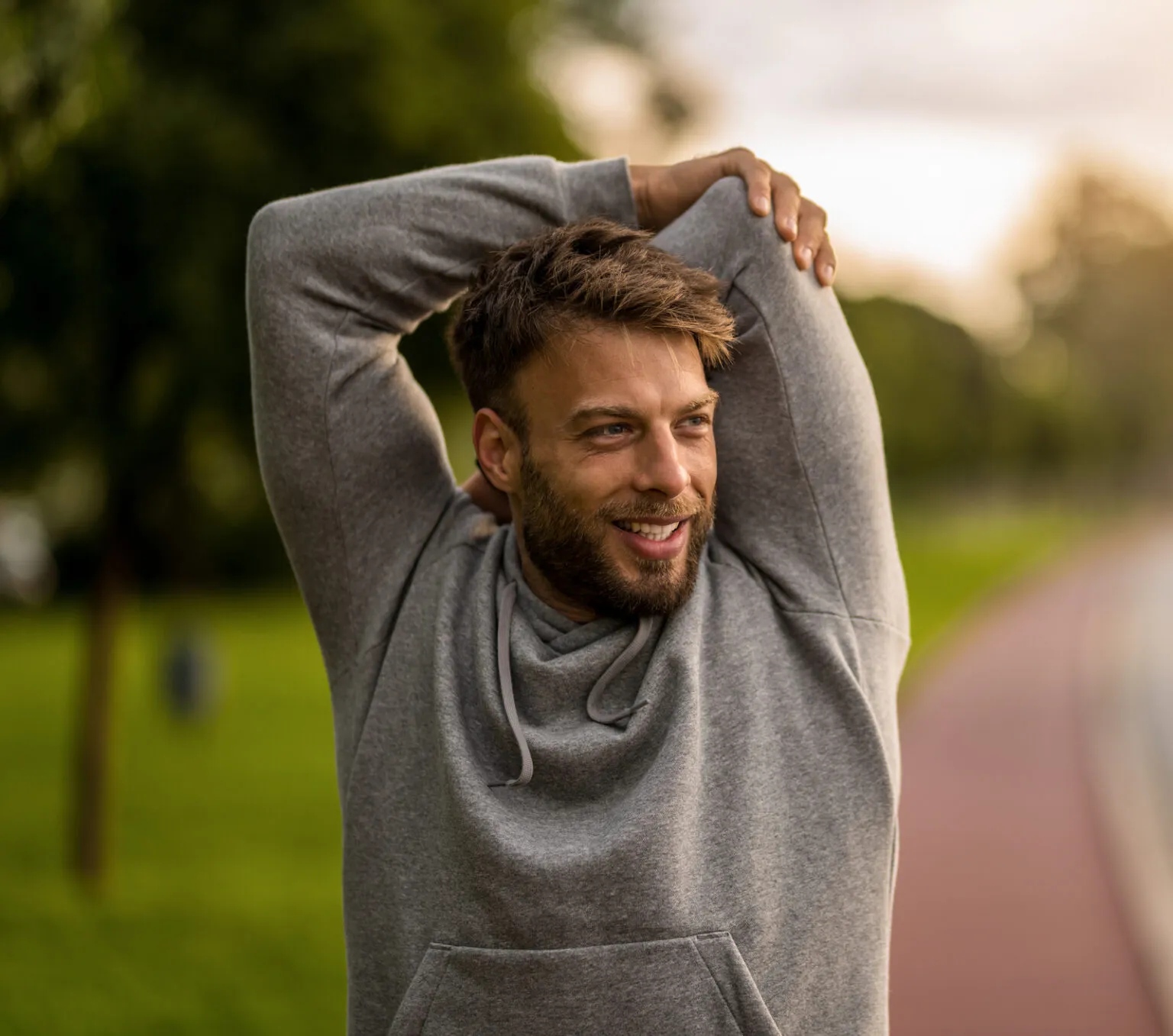 A depiction of man, doing some exercise through hands in morning , having plantlife in form of trees and grass and a simple cemented path in his background.